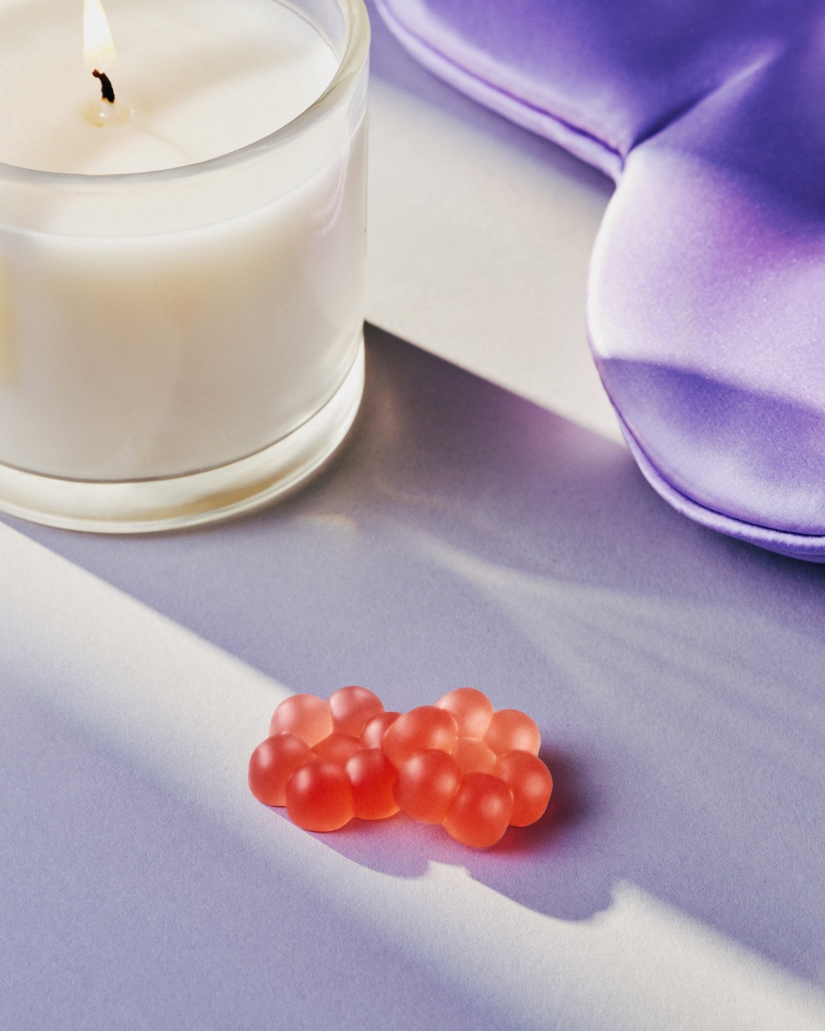 A styled product shot featuring a lit white candle in a clear glass jar, a purple satin sleep mask, and three small red boba pearls on a lavender surface with soft shadows.
