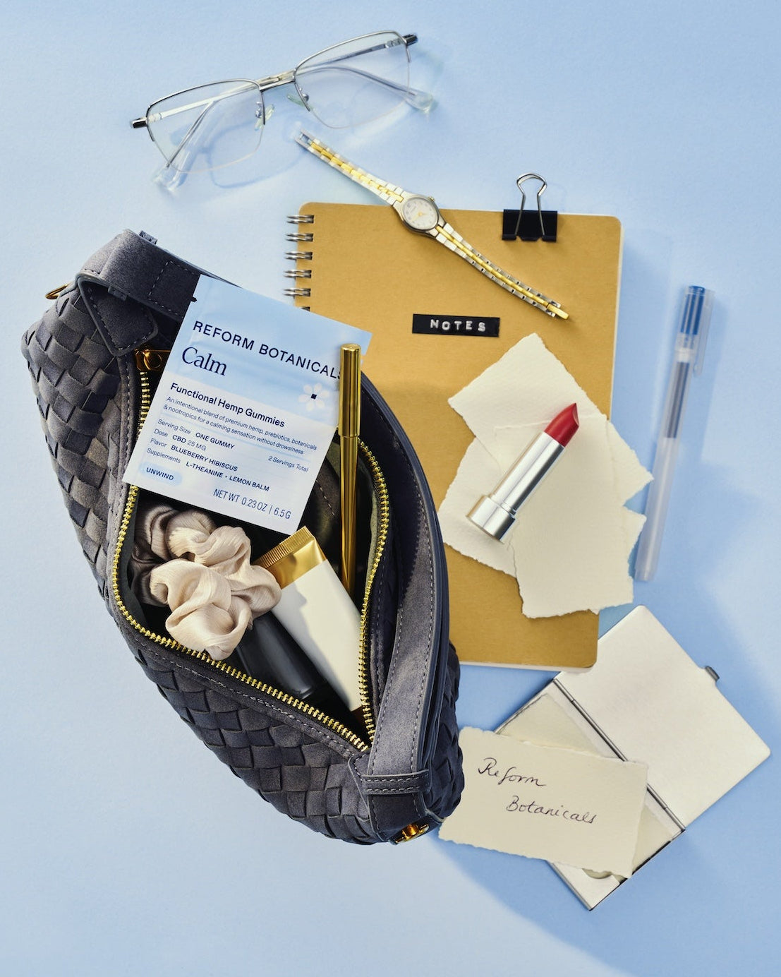 A woven black cosmetic pouch filled with skincare items, arranged on a light blue background with stationery and accessories around it.