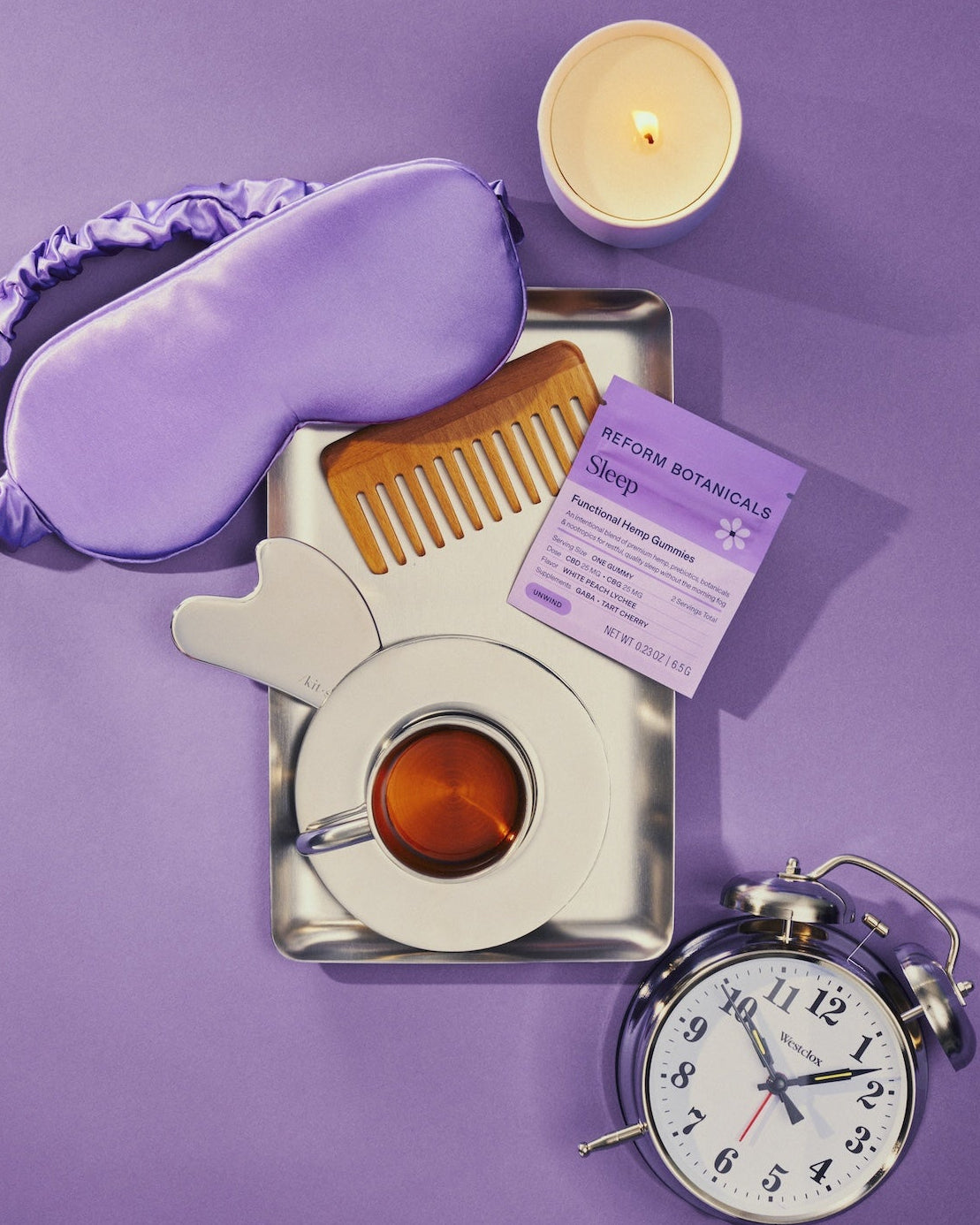 A styled flat-lay of sleep-themed items on a purple background: a lavender satin sleep mask, a small candle, a metal tray holding a wooden comb, a cup of tea, a lavender packet labeled 'KITCHEN BOTANICS Sleep', and a silver alarm clock.